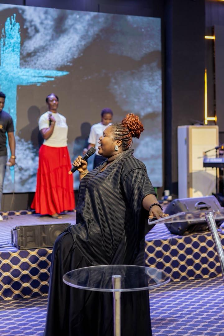 Mercy Sinclear in black singing on stage with vocalists and a cross on the screen behind