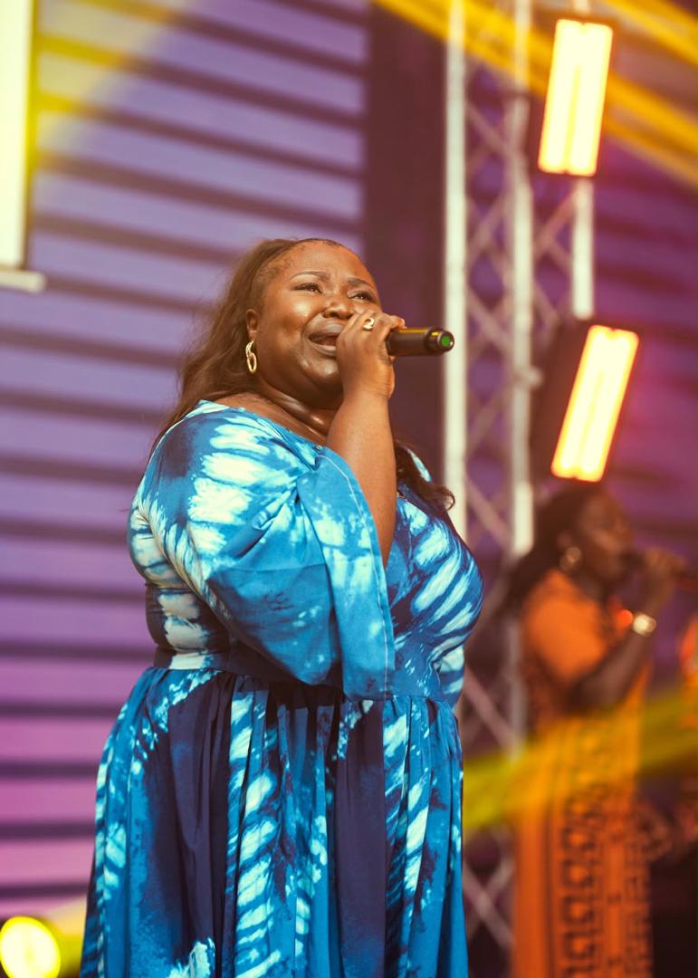 Mercy Sinclear singing on stage in a blue tie-dye dress under purple and gold lights