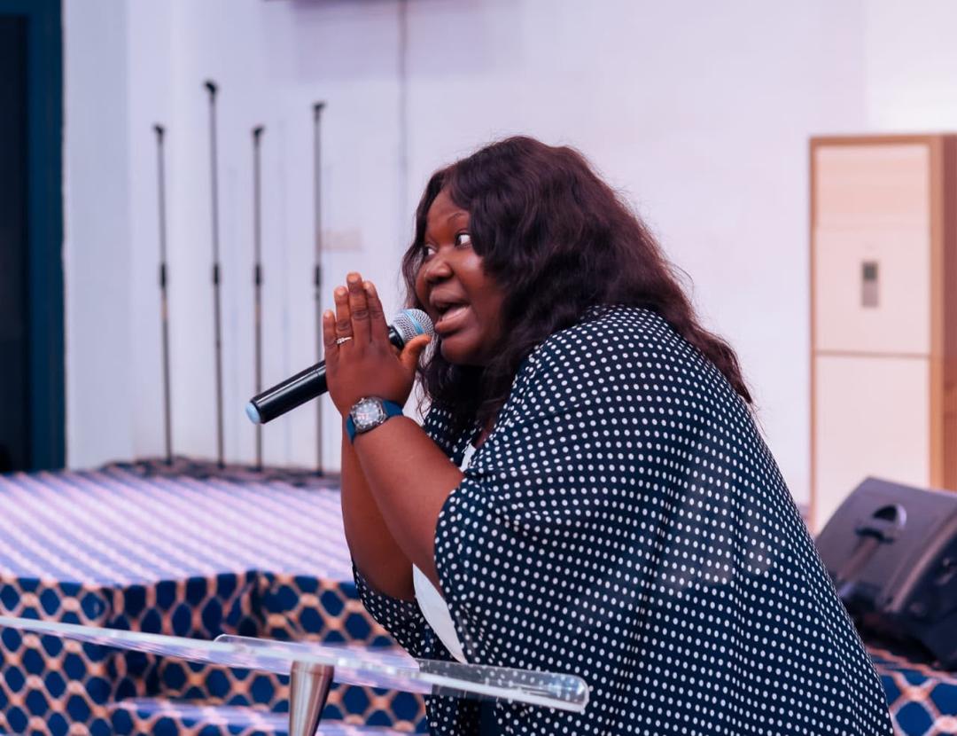 Mercy Sinclear in a polka-dot outfit speaking into a microphone, hand raised